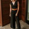 Full body view of model wearing the Nova Black Sleeveless Belted Jumpsuit which features black knit fabric, upper pleated details, wide pant legs, v-neckline, sleeveless, monochrome back zipper with hook closure and an adjustable camel brown belt with a g