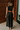 Full body back view of model wearing the Nova Black Sleeveless Belted Jumpsuit which features black knit fabric, upper pleated details, wide pant legs, v-neckline, sleeveless, monochrome back zipper with hook closure and an adjustable camel brown belt wit