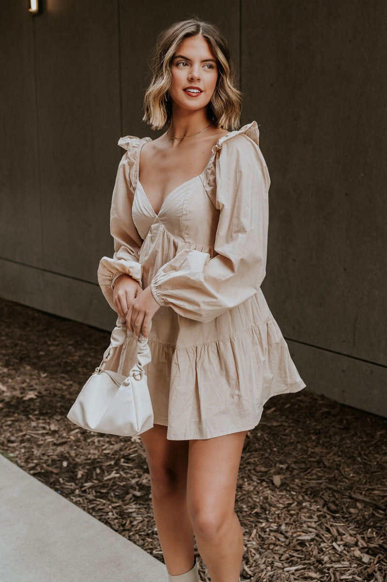 Front view of model wearing the Stella Beige Long Sleeve Mini Dress which features beige lightweight fabric, mini length, flared skirt, beige lining, sweetheart neckline, ruffle details, long sleeves with elastic wrists and smocked back.