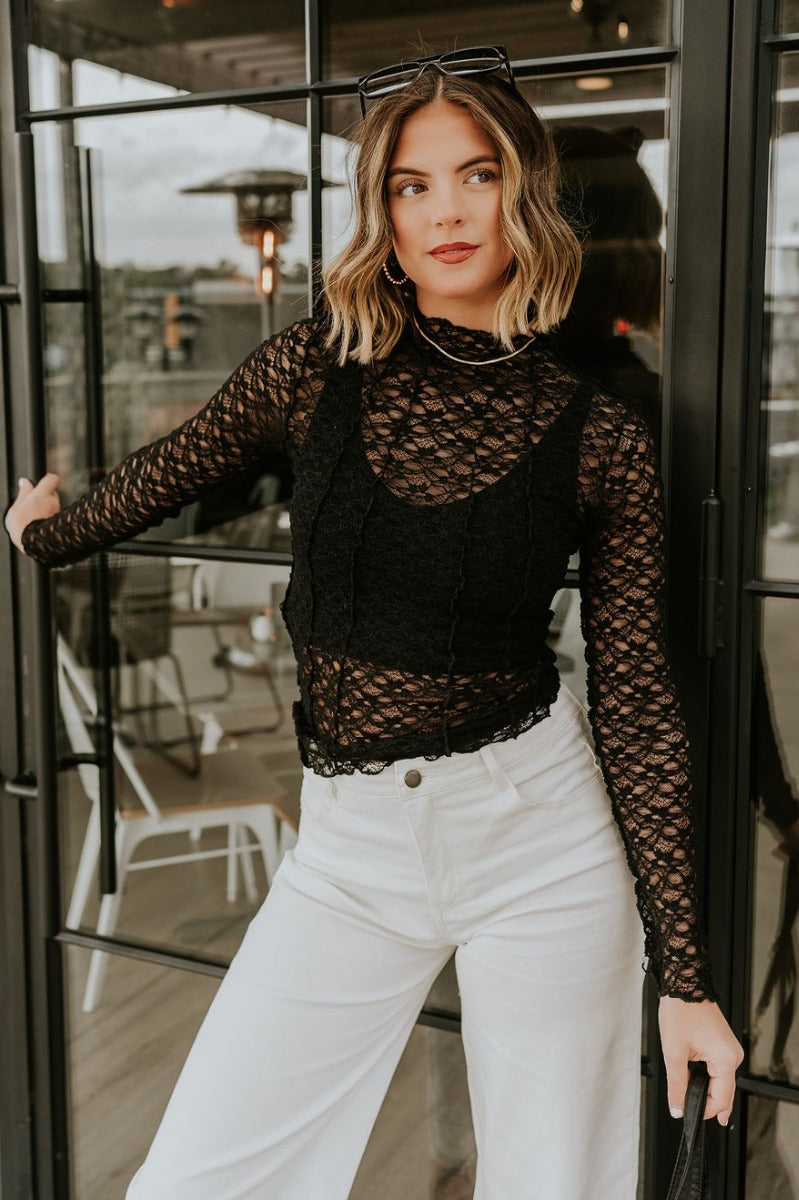front view of model wearing the Abby Black Lace Long Sleeve Top that has black sheer fabric, monochrome lace floral pattern, lettuce hem details, a high neck and long sleeves.