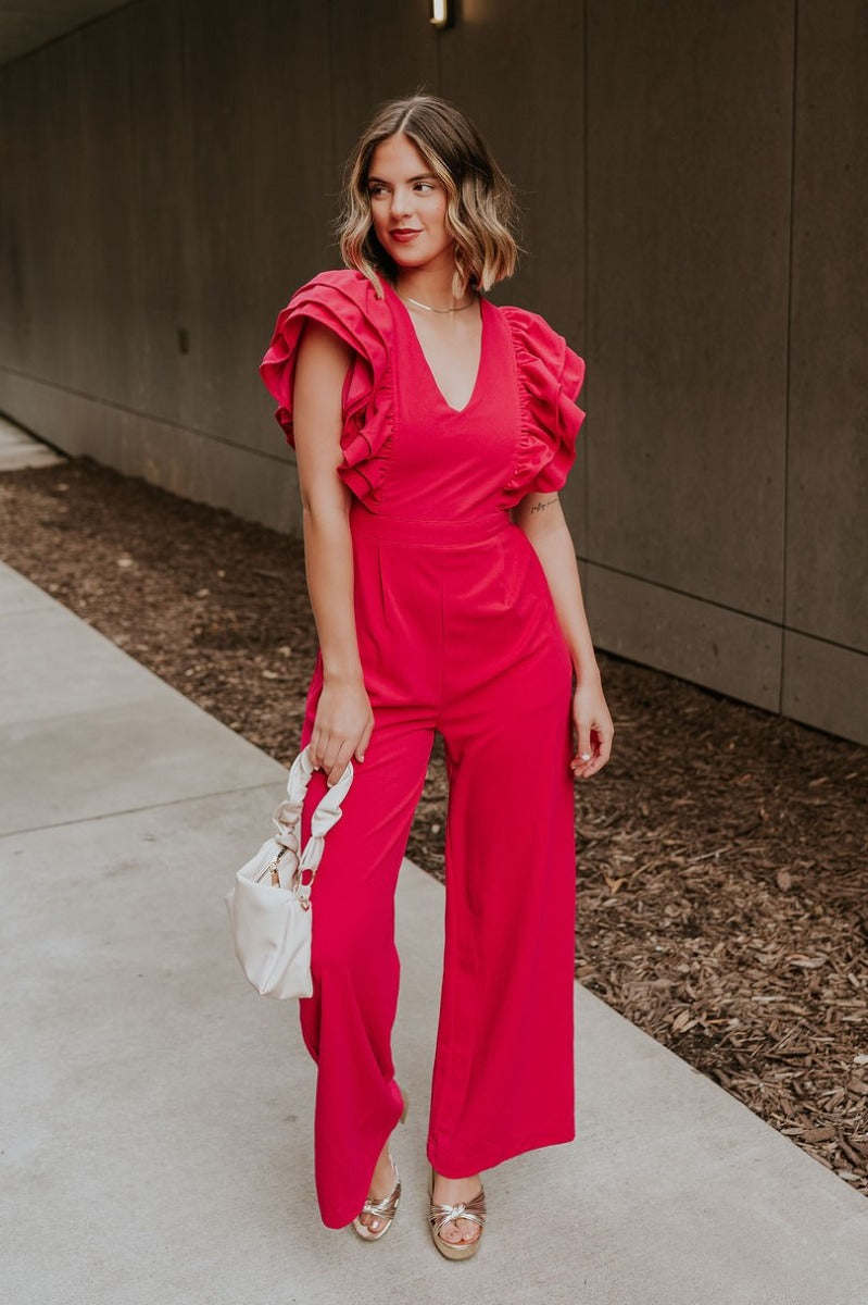 Full body front view of model wearing the Giana Hot Pink Wide Leg Ruffle Jumpsuit that has hot pink knit fabric, pockets, a ruffle details, a v-neck, a back zipper, and flare pant legs.