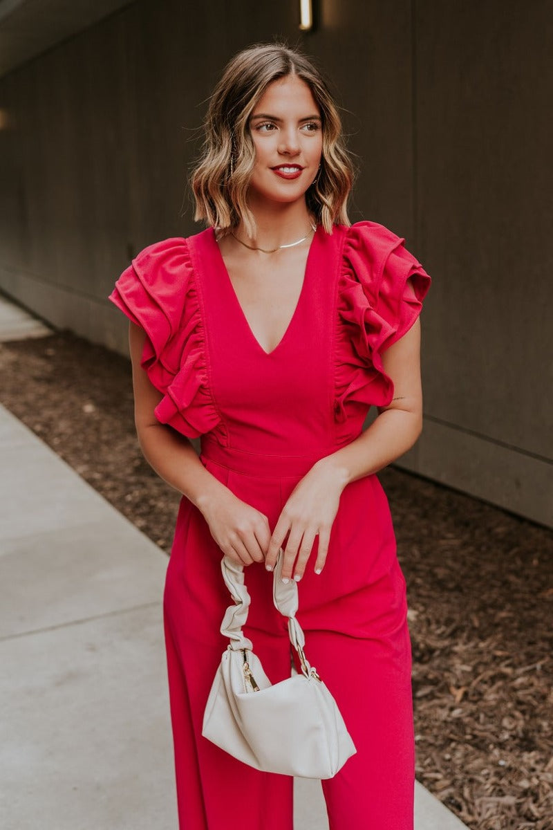 Upper front view of model wearing the Giana Hot Pink Wide Leg Ruffle Jumpsuit that has hot pink knit fabric, pockets, a ruffle details, a v-neck, a back zipper, and flare pant legs.