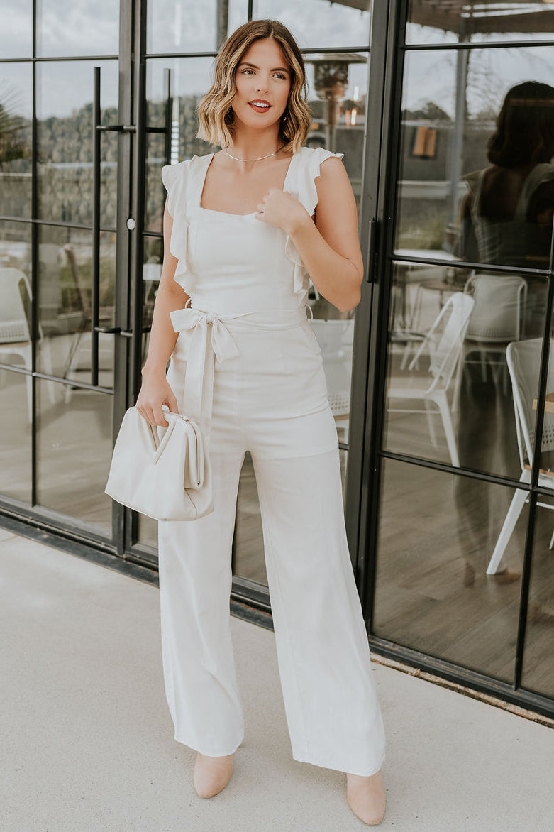 Full body view of model wearing the Clara Cream Ruffle Sleeveless Jumpsuit which features cream knit fabric, two slit pockets, waist tie closure with belt loops, square neckline, ruffle straps, sleeveless and monochrome back zipper with hook closure.