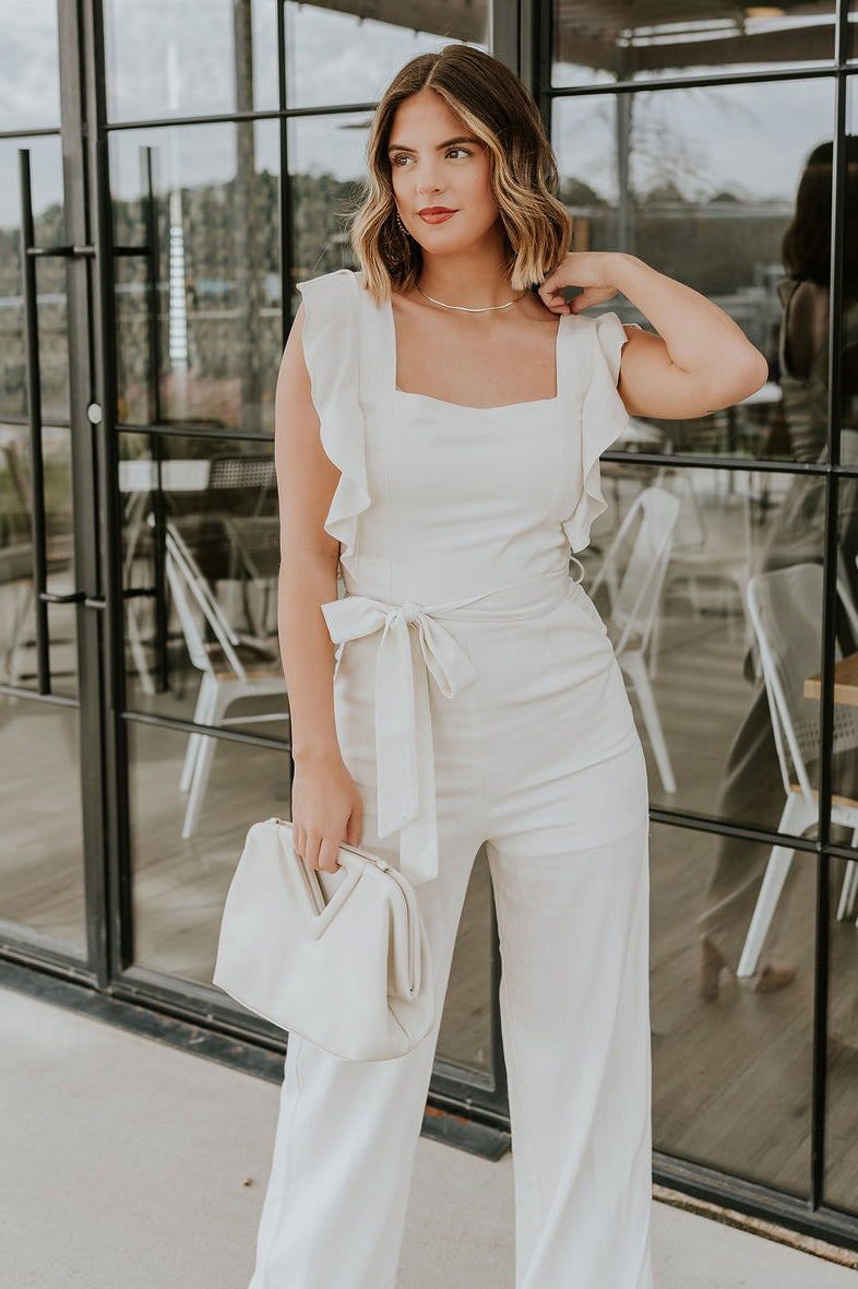 Front view of model wearing the Clara Cream Ruffle Sleeveless Jumpsuit which features cream knit fabric, two slit pockets, waist tie closure with belt loops, square neckline, ruffle straps, sleeveless and monochrome back zipper with hook closure.