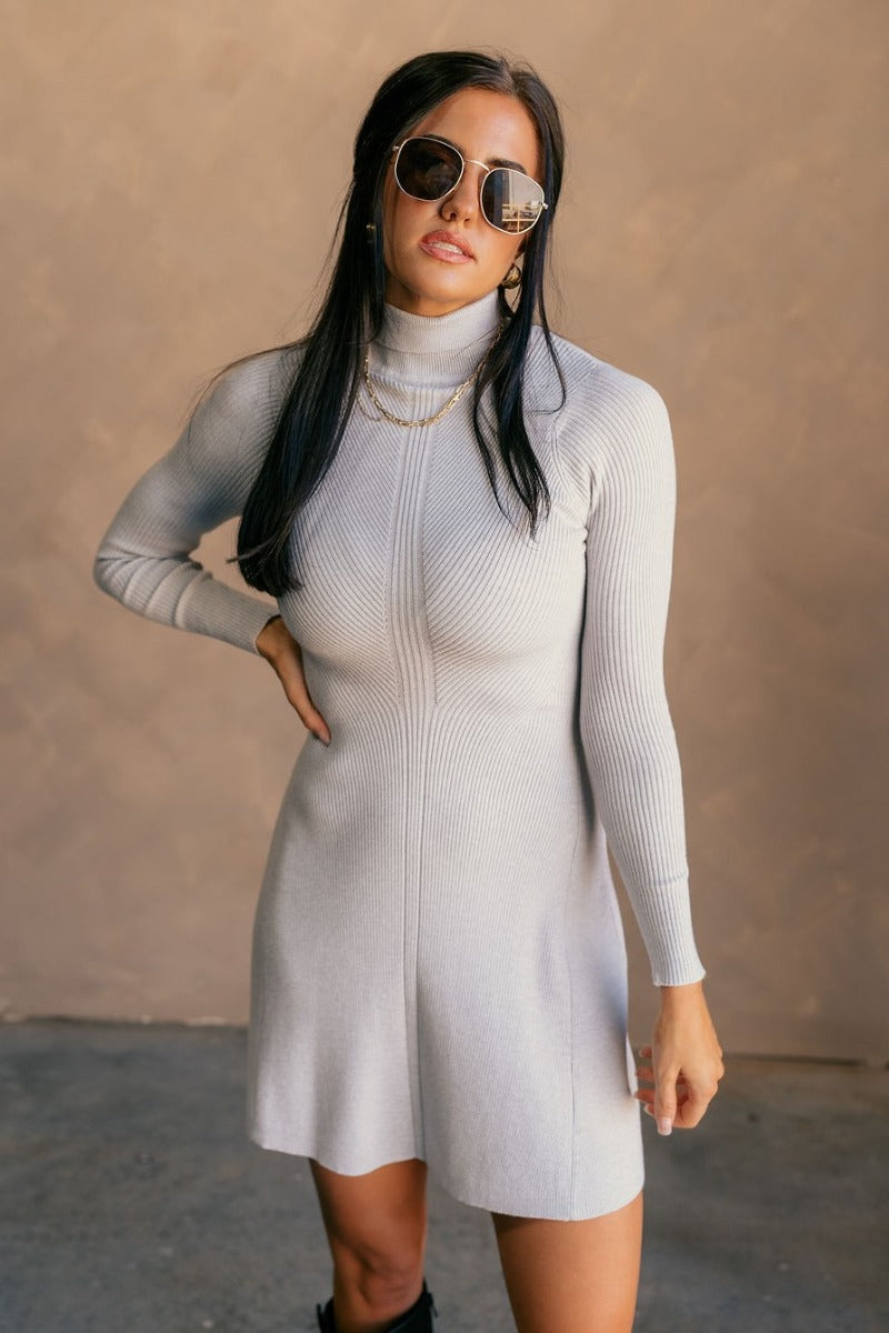 Front view of model wearing the Melina Grey Turtleneck Sweater Dress that has light grey ribbed knit fabric, a flared skirt, mini length, a turtleneck neckline, and long sleeves.