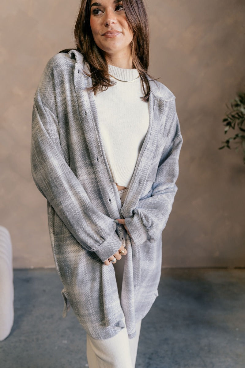 Front view of model wearing the Clara Grey Plaid Knit Long Sleeve Shacket which features grey and white knit fabric, a geometric plaid pattern, mid thigh length hem, two pockets on each side, tortoise buttons, a collared neckline, a left front chest pocke