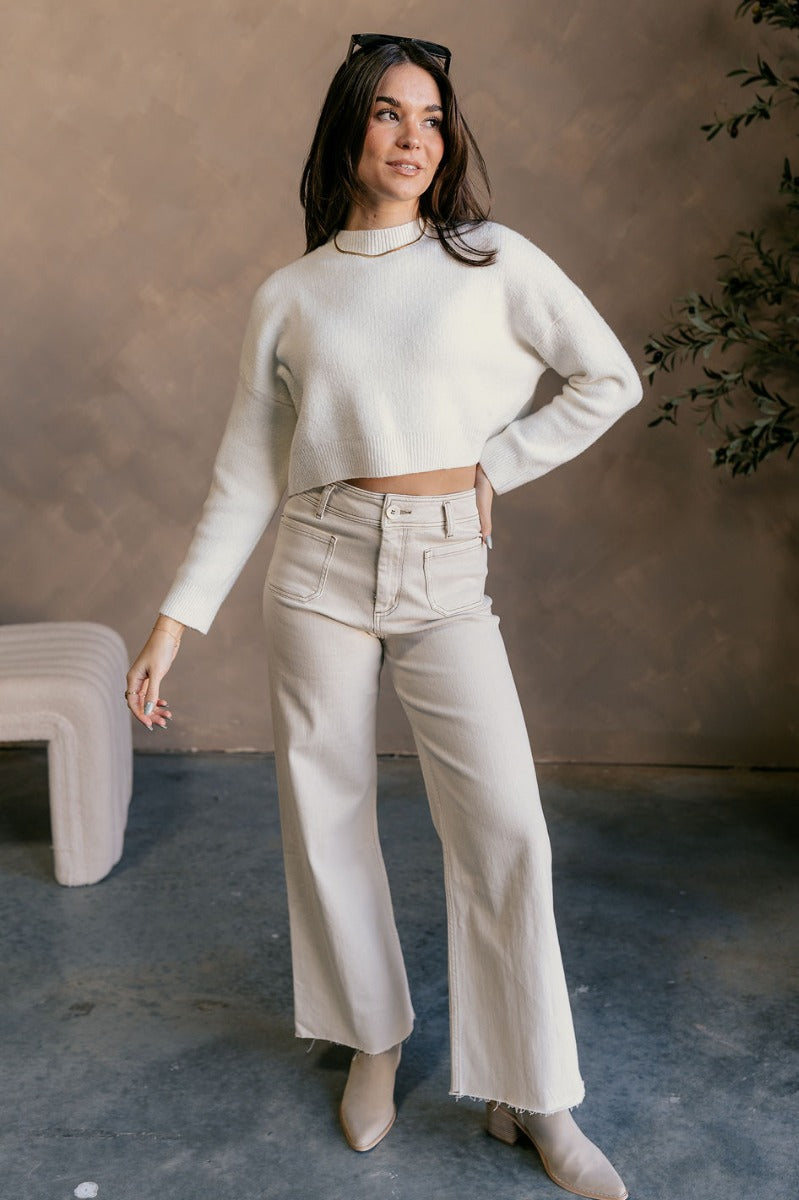 Full body view of model wearing the Emily Ivory Cropped Long Sleeve Sweater which features ivory knit fabric, a ribbed hem, a cropped waist, a slight high neckline, dropped shoulders, and long sleeves.