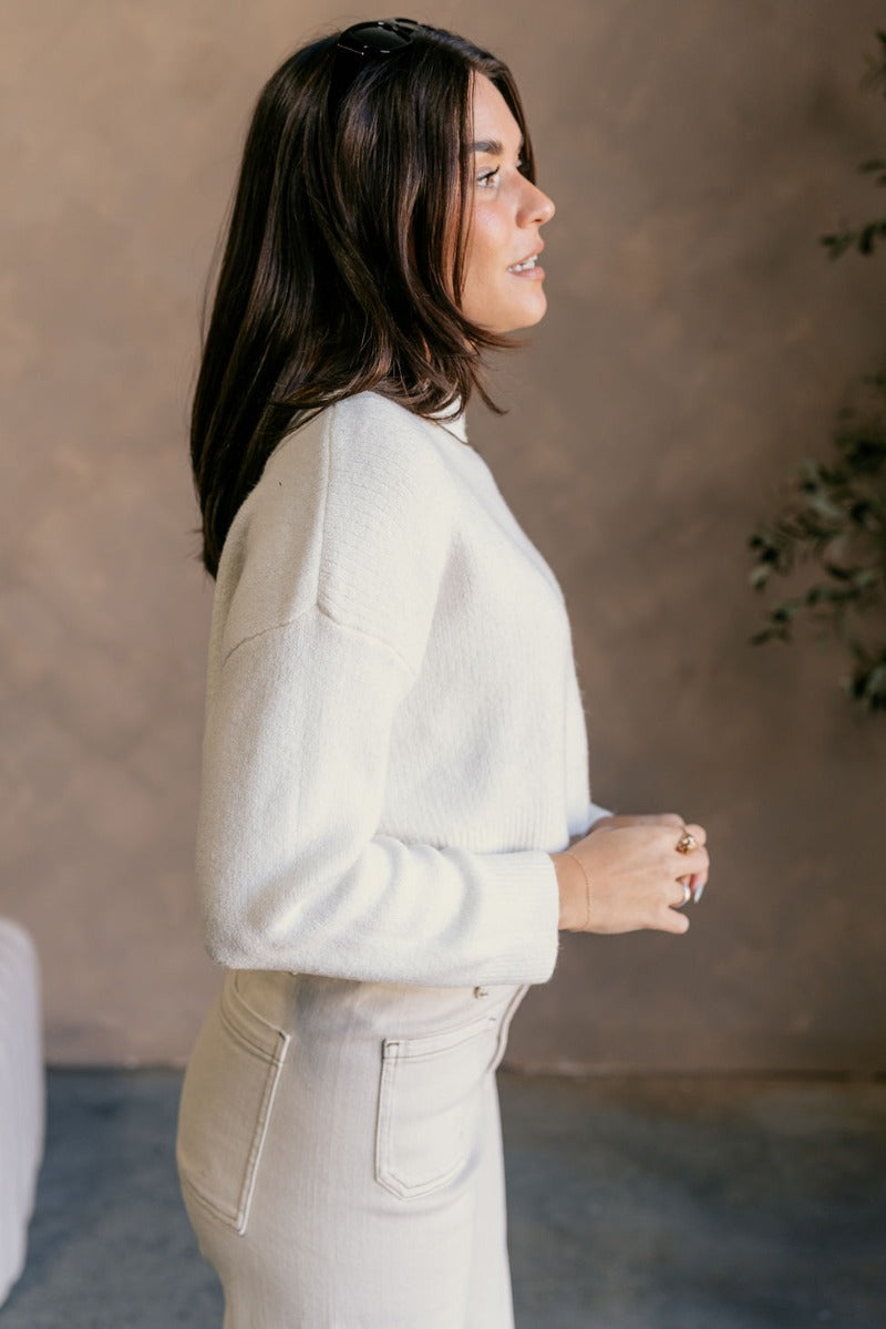 Side view of model wearing the Emily Ivory Cropped Long Sleeve Sweater which features ivory knit fabric, a ribbed hem, a cropped waist, a slight high neckline, dropped shoulders, and long sleeves.