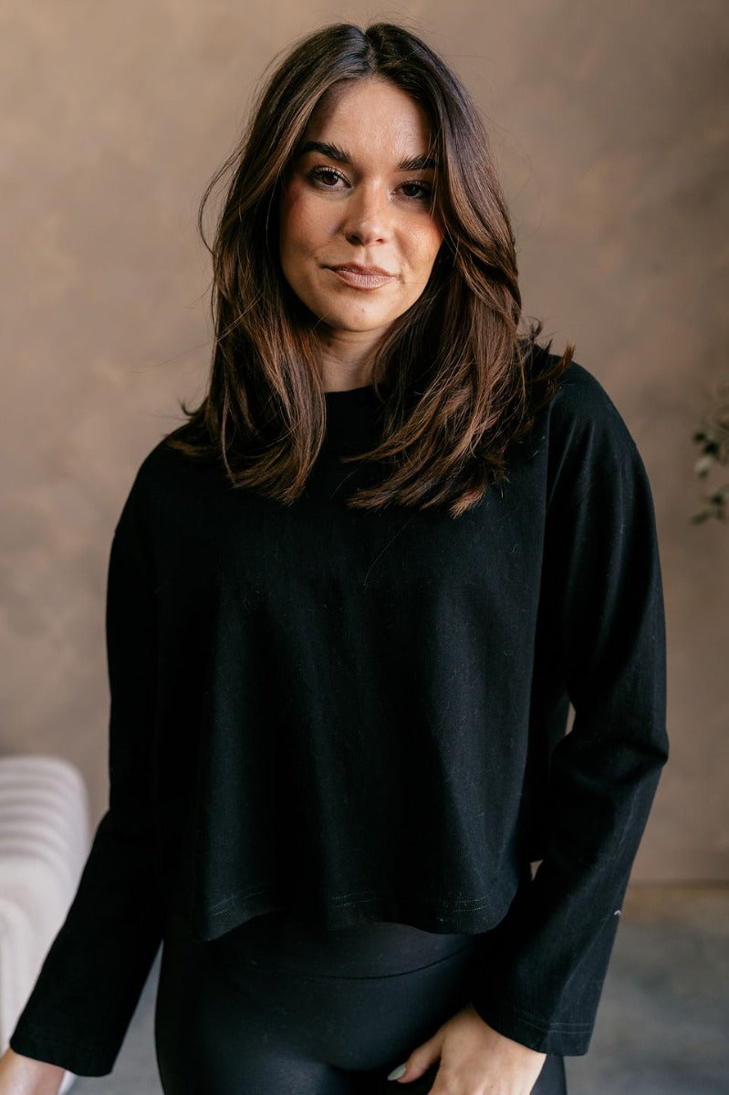 Front view of model wearing the Nicole Black Cropped Long Sleeve Top which features black cotton fabric, a cropped waist, a round neckline, and long sleeves.