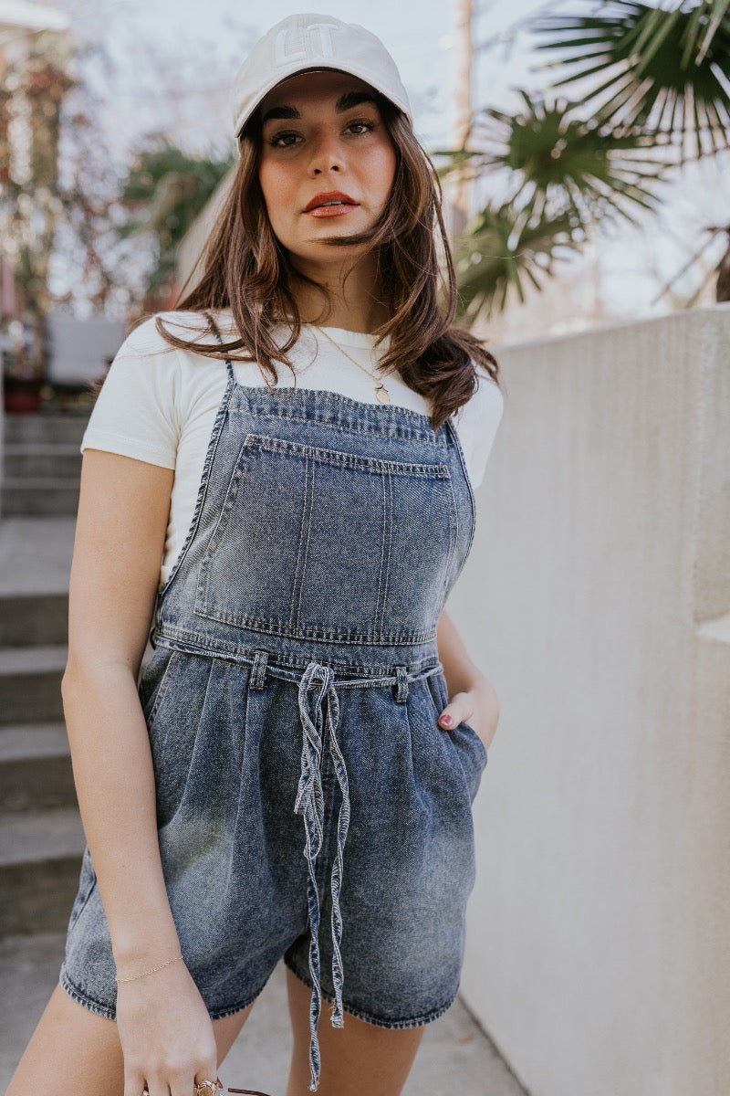 Front detail image of model wearing The Marina Washed Denim Romper features medium wash denim fabric, two front pockets, two back pockets, belt loops with a drawstring tie, one front chest pocket, a halter neckline, adjustable tie straps, a side zipper cl
