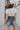 Back view of model wearing The Marina Washed Denim Romper features medium wash denim fabric, two front pockets, two back pockets, belt loops with a drawstring tie, one front chest pocket, a halter neckline, adjustable tie straps, a side zipper closure and