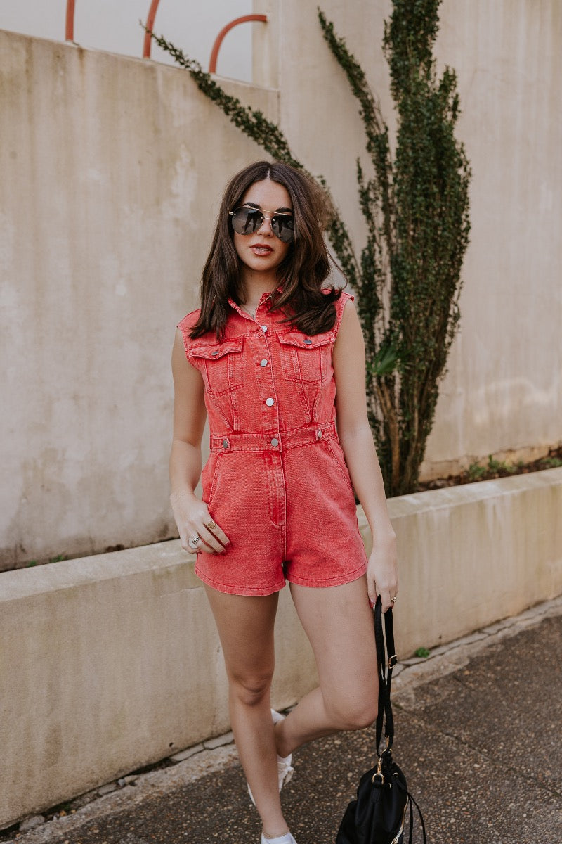 front full body image of modeal wearing The Emily Washed Red Denim Sleeveless Romper features washed red denim fabric, two front pockets, silver button details, button-up front, two front chest buttoned pockets, two back pockets, a collared neckline and a