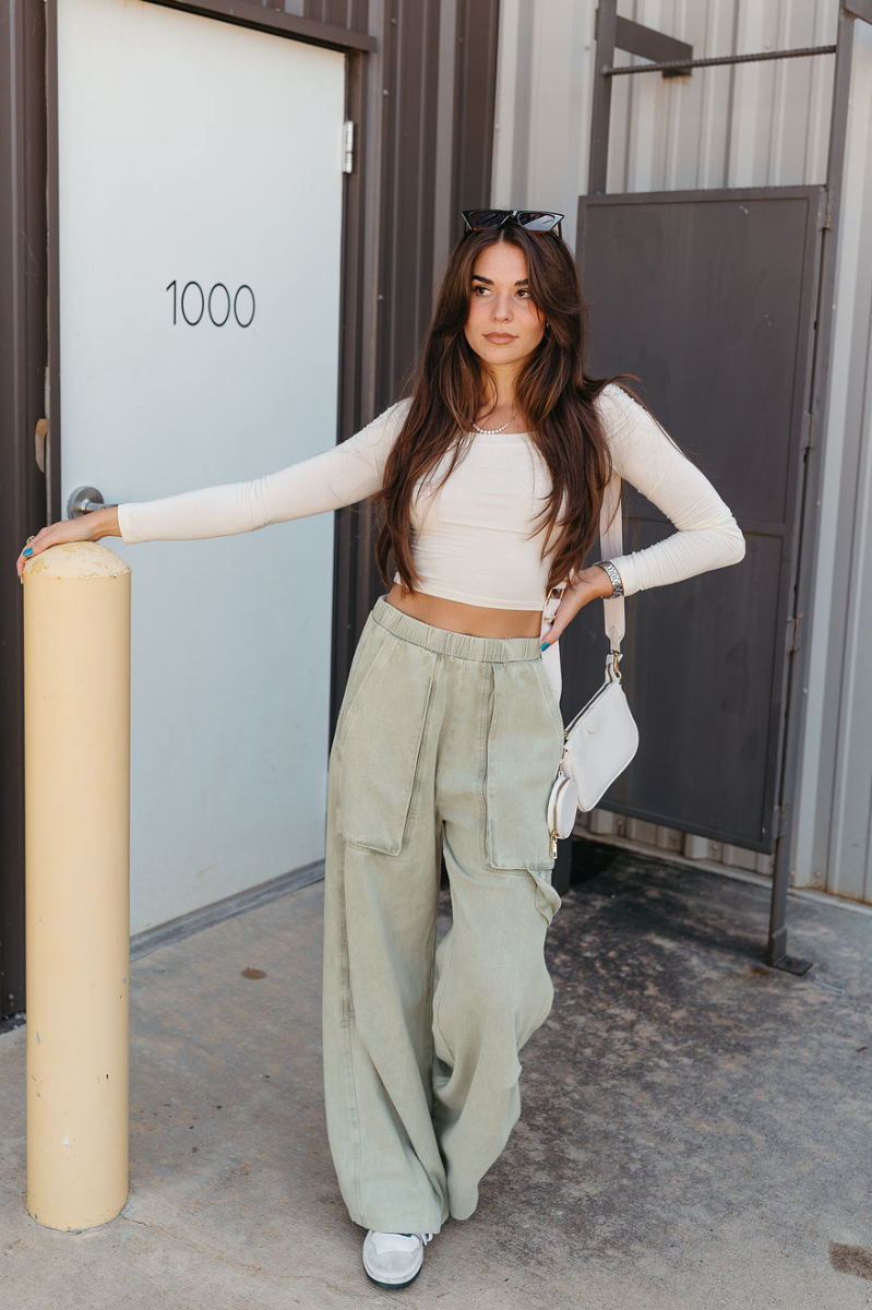 Full body view of model wearing the Ellison Denim Cargo Pants in Light Sage which features light olive cotton fabric, two front pockets, a side cargo loop detail, an elastic waistband and wide legs.