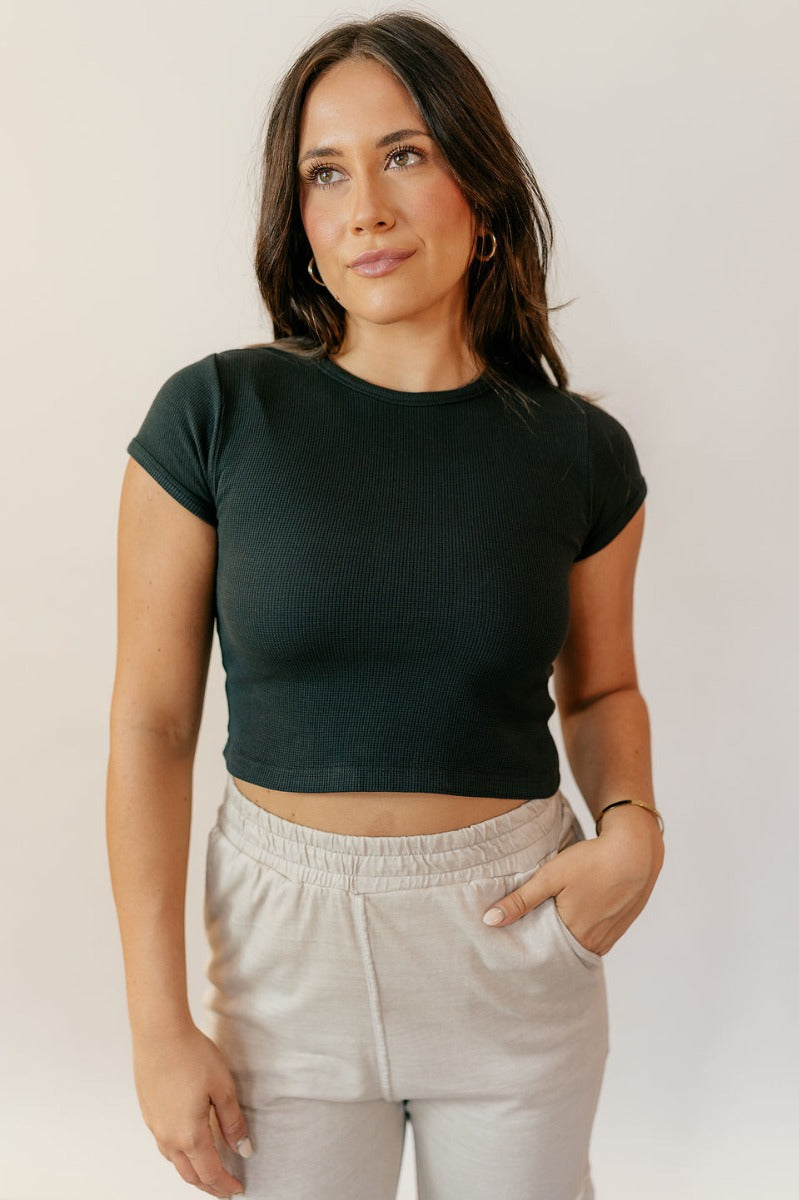 Front view of model wearing the Taryn Black Basic Short Sleeve Top which features black textured cotton fabric, a cropped waist, a round neckline and short sleeves.