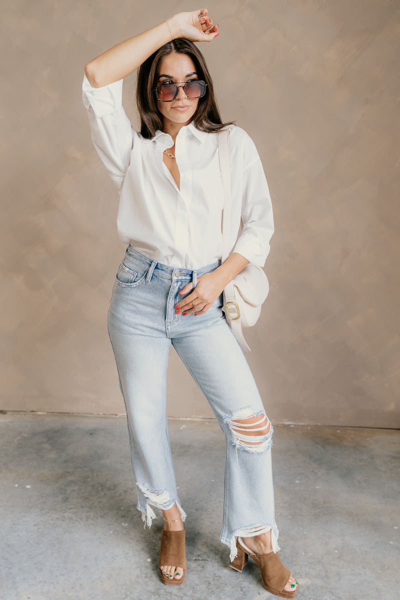 Full body view of model wearing the Gia White Button-Up Collared Long Sleeve Top which features white cotton fabric, monochromatic buttons, a collared neckline, and long sleeves with buttoned cuffs.