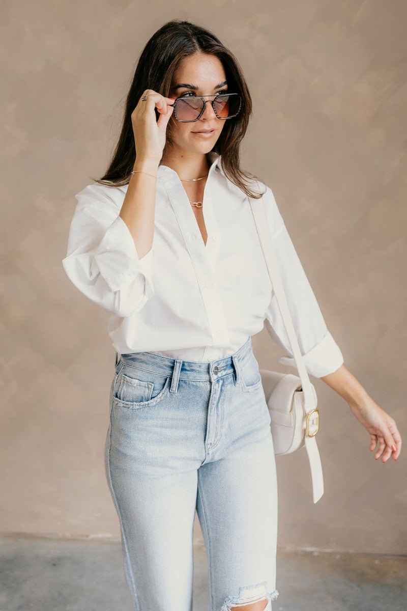Front view of model wearing the Gia White Button-Up Collared Long Sleeve Top which features white cotton fabric, monochromatic buttons, a collared neckline, and long sleeves with buttoned cuffs.