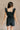 Back view of model wearing the Avril Black Denim Ruffle Sleeveless Romper which features black washed denim fabric, pockets on each side, a front zip up, an elastic waistband, a square neckline, and ruffle straps.