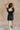 Full body back view of model wearing the Avril Black Denim Ruffle Sleeveless Romper which features black washed denim fabric, pockets on each side, a front zip up, an elastic waistband, a square neckline, and ruffle straps.