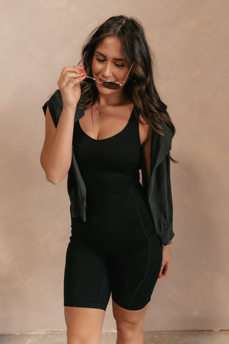 Front view of model wearing the Amara Black Athletic Onesie Bodysuit that has black atheisure fabric, side pockets, built-in-padding, monochrome stitching, and thick straps. Sweater is draped on model's shoulders.