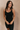 Front view of model wearing the Amara Black Athletic Onesie Bodysuit that has black atheisure fabric, side pockets, built-in-padding, monochrome stitching, and thick straps. Sweater is draped on model's shoulders.