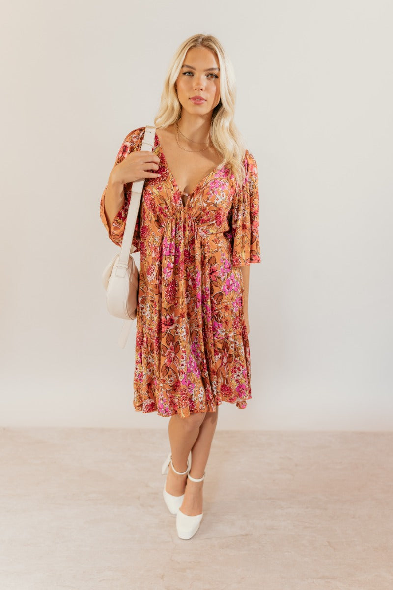 Full body front view of model wearing the Rachel Rust Floral Short Sleeve Mini Dress that has rust fabric with a floral pattern, mini length, flared skirt, a plunge neck, an open back with a tie , and short flare sleeves.