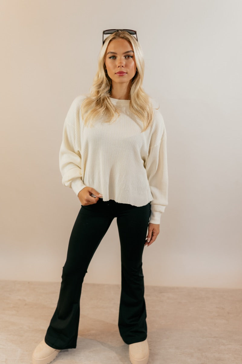 Full body front view of model wearing the Ada Cream Waffle-Knit Long Sleeve Top that features cream waffle knit fabric, a raw hem, a round neckline, dropped shoulders, and long sleeves with cuffs.