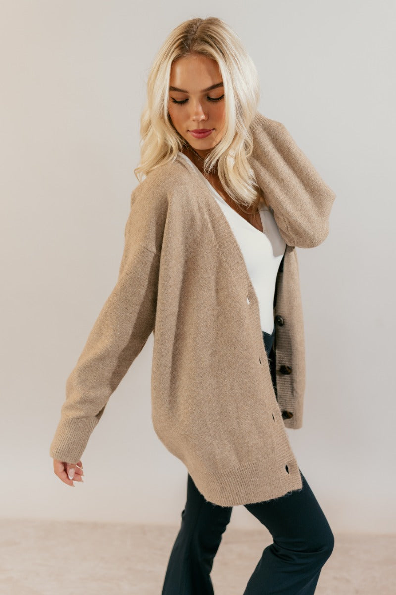 Side view of model wearing the Oaklynn Mocha Oversized Cardigan that has mocha brown knit fabric, ribbed hem, a button-up front with dark tortoise buttons, a v-neckline, and long sleeves with cuffs.