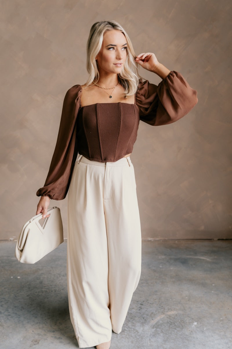 Full body front view of model wearing the Andrea Brown Long Sleeve Cropped Corset Top that has brown knit fabric, a cropped waist, boning details, a square neckline, and sheer puff sleeves with elastic wrists.