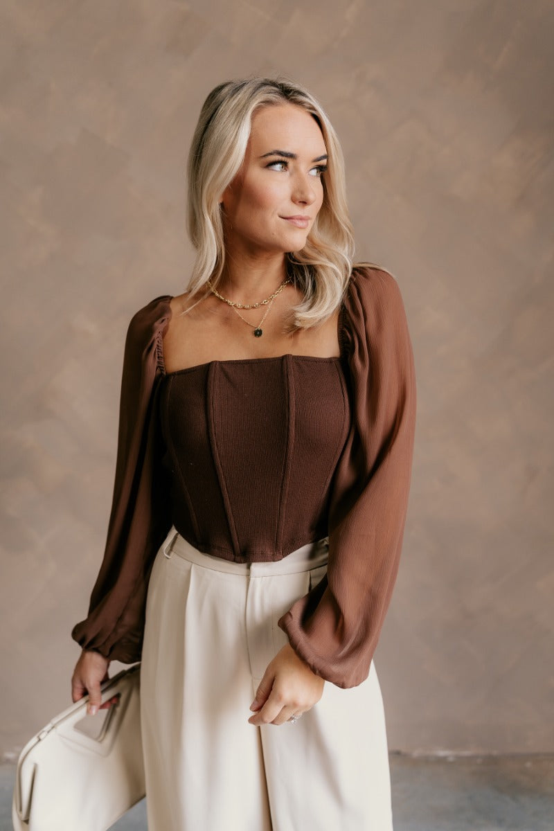 front view of model wearing the Andrea Brown Long Sleeve Cropped Corset Top that has brown knit fabric, a cropped waist, boning details, a square neckline, and sheer puff sleeves with elastic wrists.