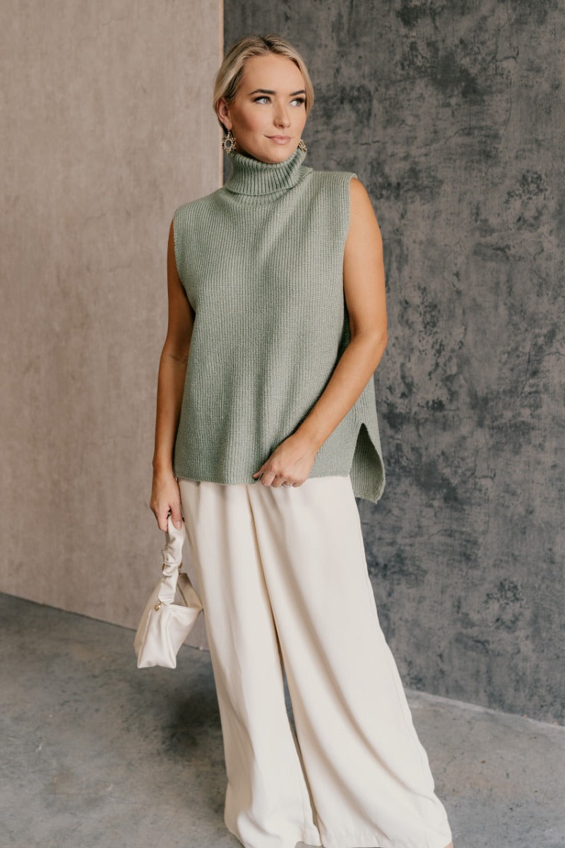 Full body front view of model wearing the Wren Sage Sleeveless Turtleneck Sweater that features sage green knit fabric, a high-low hem, slits on each side, a turtleneck neckline and a sleeveless design.