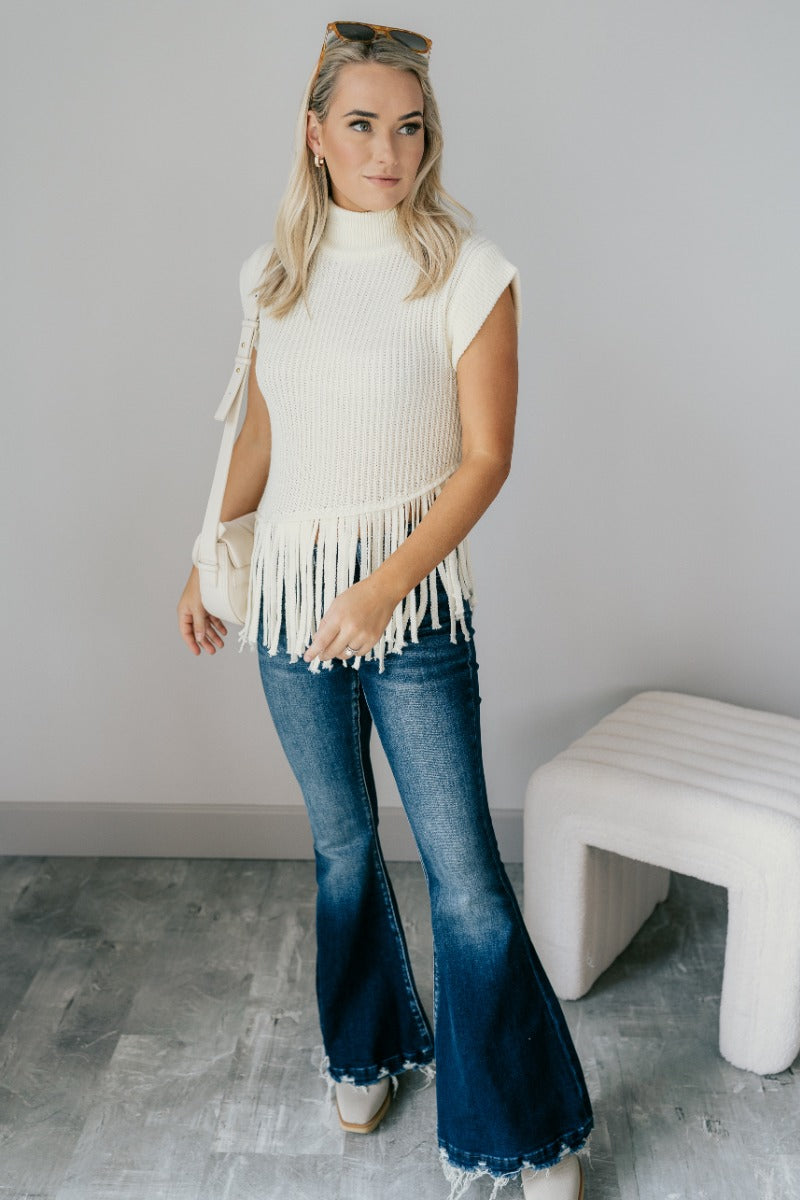 Full body view of model wearing the Brielle Cream Fringe Hem Sleeveless Knit Sweater which features cream knit fabric, a slanted hem with fringe details, a high neckline and a sleeveless design.