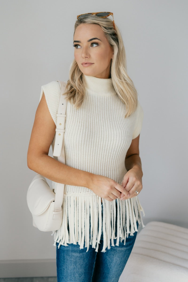 Front view of model wearing the Brielle Cream Fringe Hem Sleeveless Knit Sweater which features cream knit fabric, a slanted hem with fringe details, a high neckline and a sleeveless design.