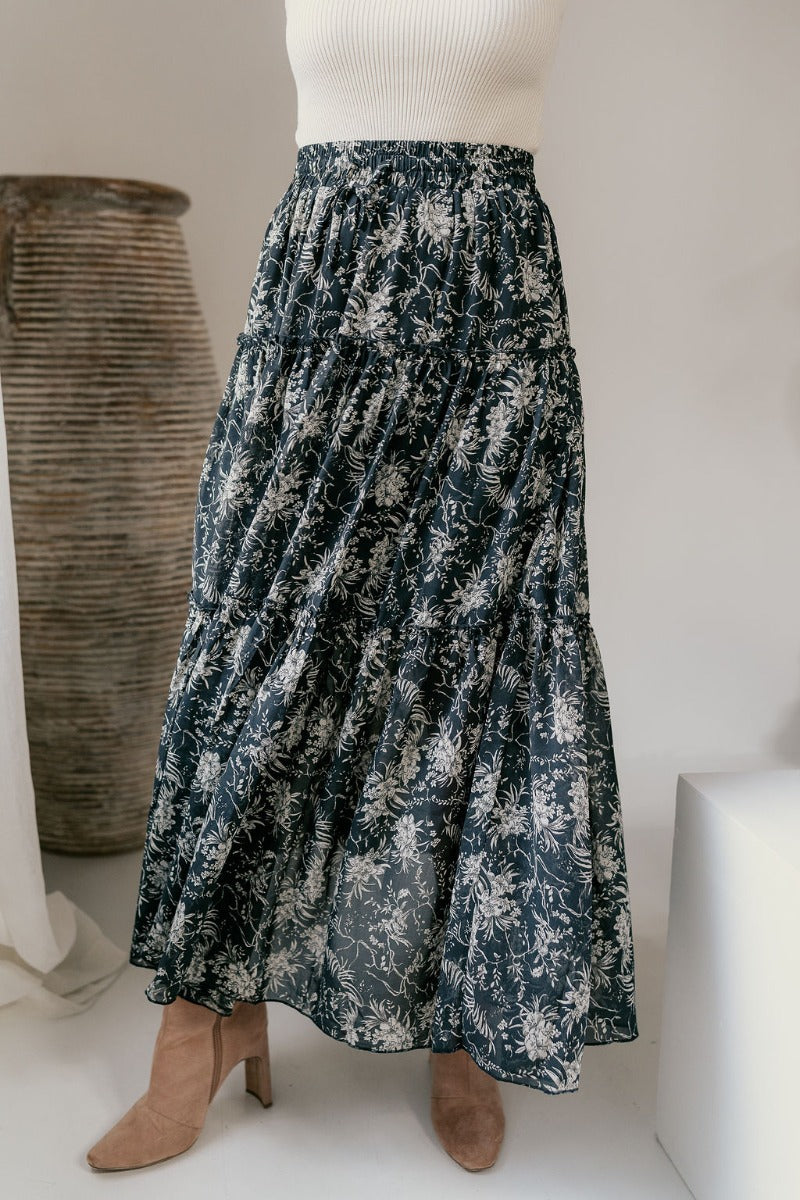 front view of model wearing the Eleanor Charcoal & White Floral Maxi Skirt that hat grey semi-sheer fabric with white floral print, maxi length, tiered ruffles, thigh-length lining, and an elastic waist.