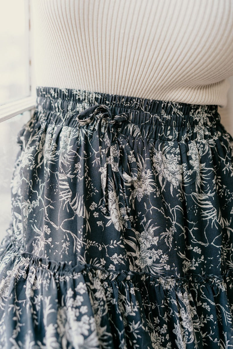 Close upper front view of model wearing the Eleanor Charcoal & White Floral Maxi Skirt that hat grey semi-sheer fabric with white floral print, maxi length, tiered ruffles, thigh-length lining, and an elastic waist.