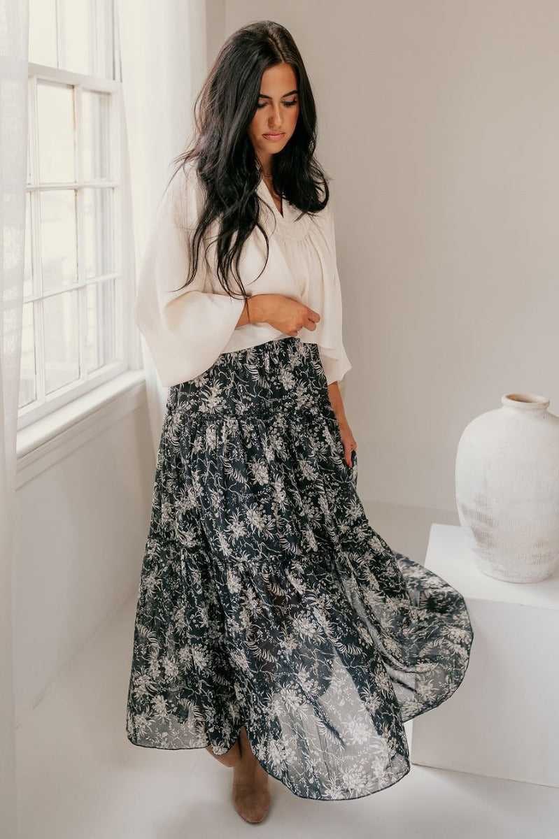 Full body front view of model wearing the Eleanor Charcoal & White Floral Maxi Skirt that hat grey semi-sheer fabric with white floral print, maxi length, tiered ruffles, thigh-length lining, and an elastic waist.