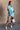 Full body view of model wearing the Delilah Washed Sky Blue Short Sleeve Romper which features washed light blue tencel fabric, two front slit pockets, an elastic waistband with a drawstring tie, a right front chest pocket, tortoise button closures, a col