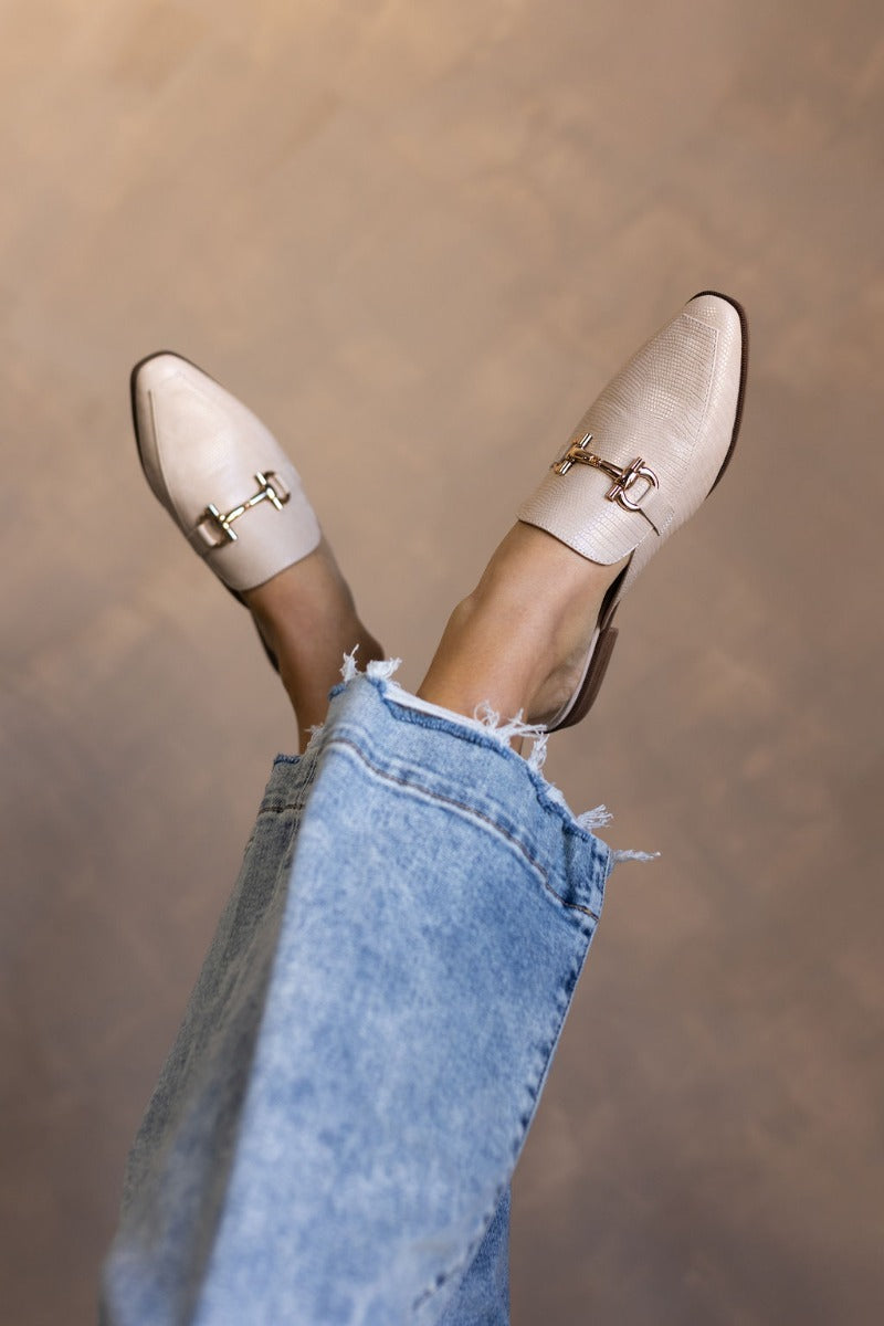 Image of model's feet while wearing the the Score Lizard Cream Gold Buckle Mule which feature cream snakeskin faux-leather, a gold buckle detail, backless slide entry, and a 1" heel.
