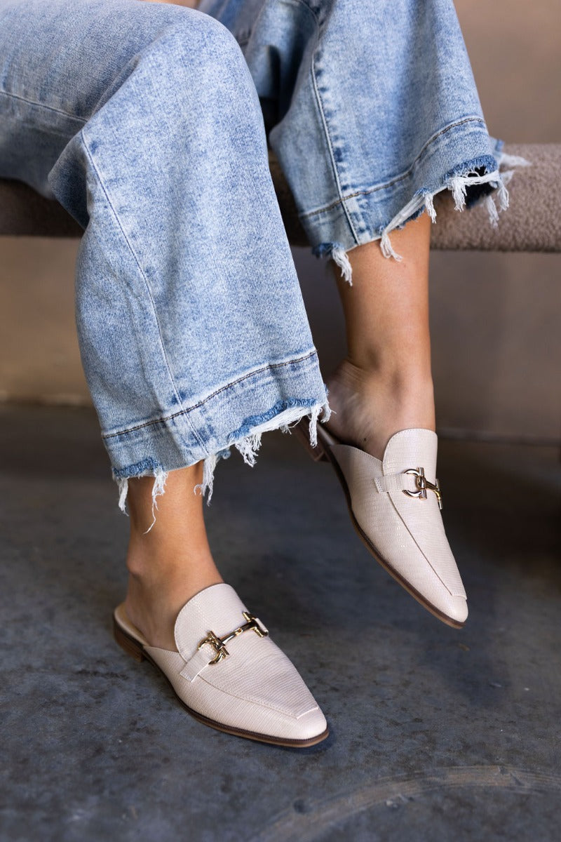 Side image of model's feet while wearing the the Score Lizard Cream Gold Buckle Mule which feature cream snakeskin faux-leather, a gold buckle detail, backless slide entry, and a 1" heel.