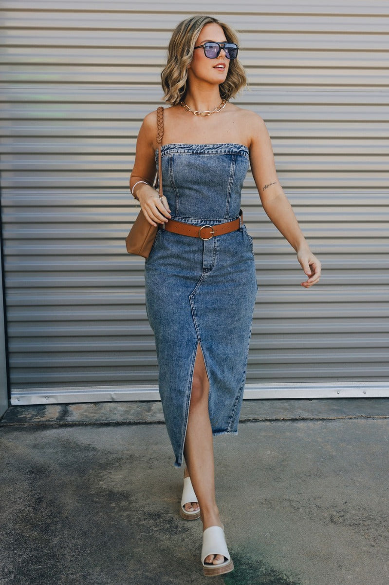 Full body view of model wearing the Carrie Washed Denim Slit Strapless Midi Dress which features medium denim wash fabric, brown stitch details, front slit detail, midi length, two front slit pockets, two back pockets, belt loops, frayed hem, straight acr