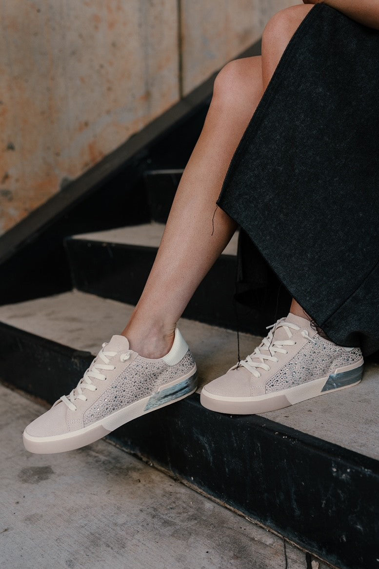 Front right view of female model wearing the Zina Crystal Sneaker in Ivory Suede which features ivory suede upper fabric, crystal stone design, white lace up closure, silver and neutral tones on sole and round toe.