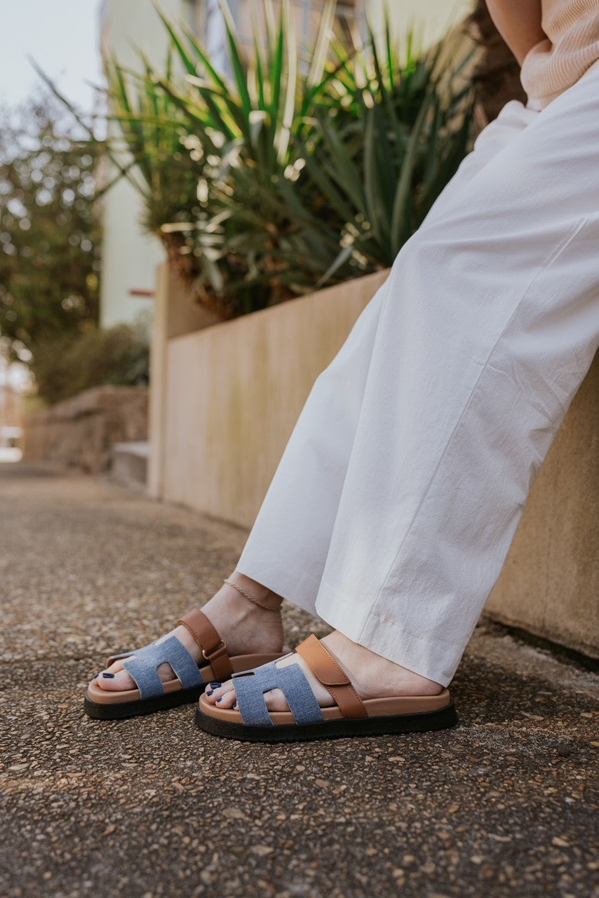Ariel view of the model holding the Mayven Sandal in Denim Multi which features a brown leather fabric cutout strappy upper with hook and loop closure, denim "h" shaped strap, round toe, black sole and slide on style.