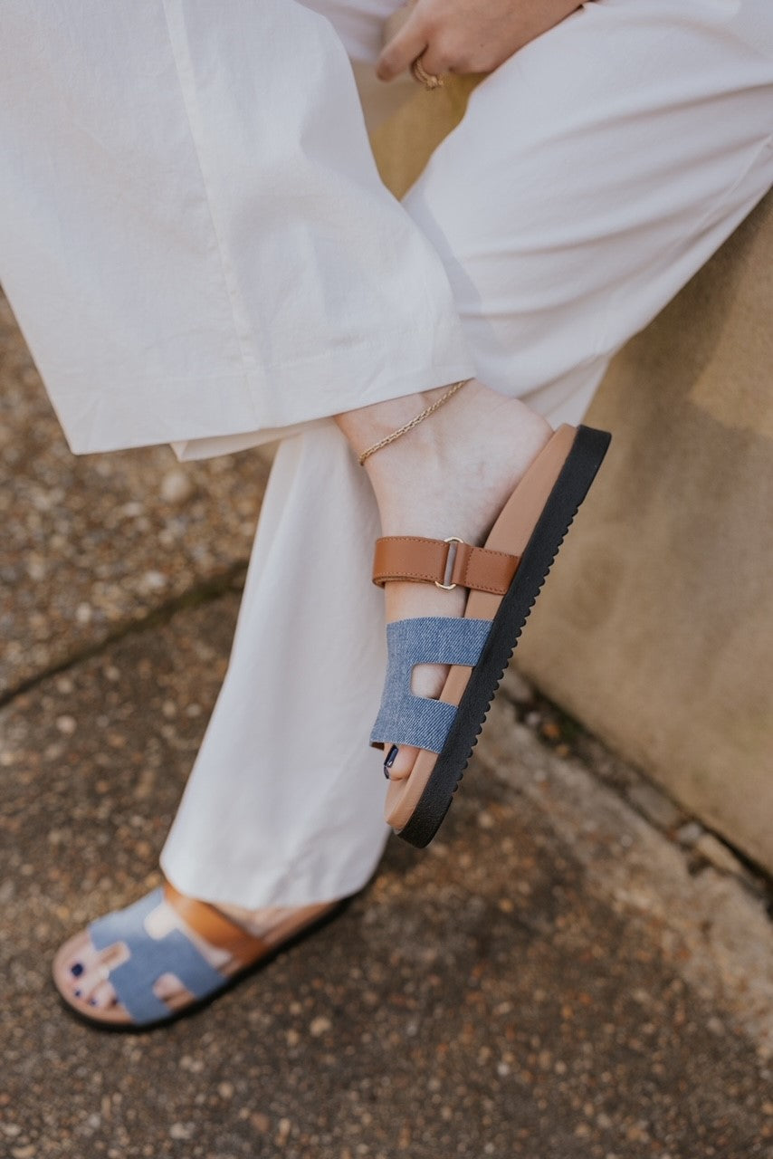 Side view of the model holding the Mayven Sandal in Denim Multi which features a brown leather fabric cutout strappy upper with hook and loop closure, denim "h" shaped strap, round toe, black sole and slide on style.