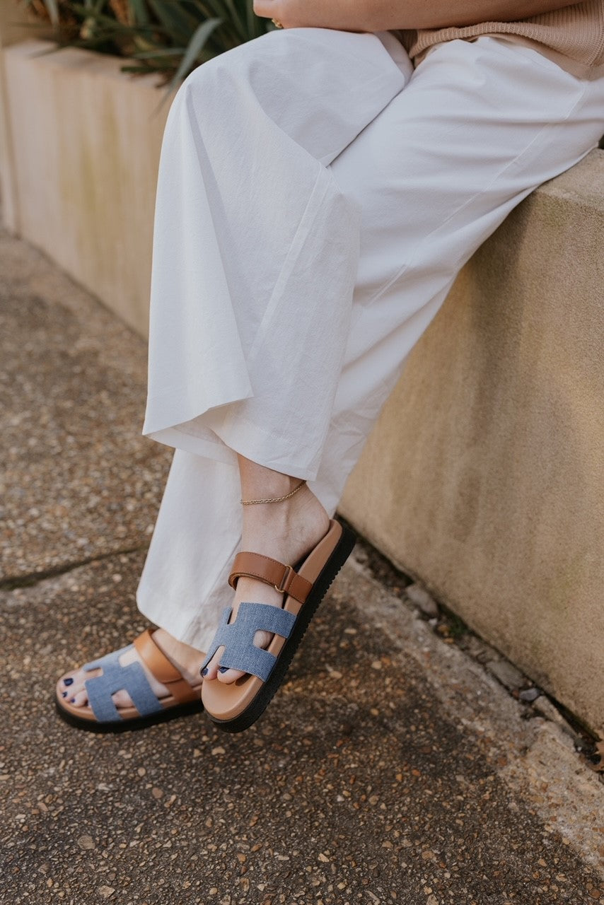 Side view of model wearing the Mayven Sandal in Denim Multi which features a brown leather fabric cutout strappy upper with hook and loop closure, denim "h" shaped strap, round toe, black sole and slide on style.
