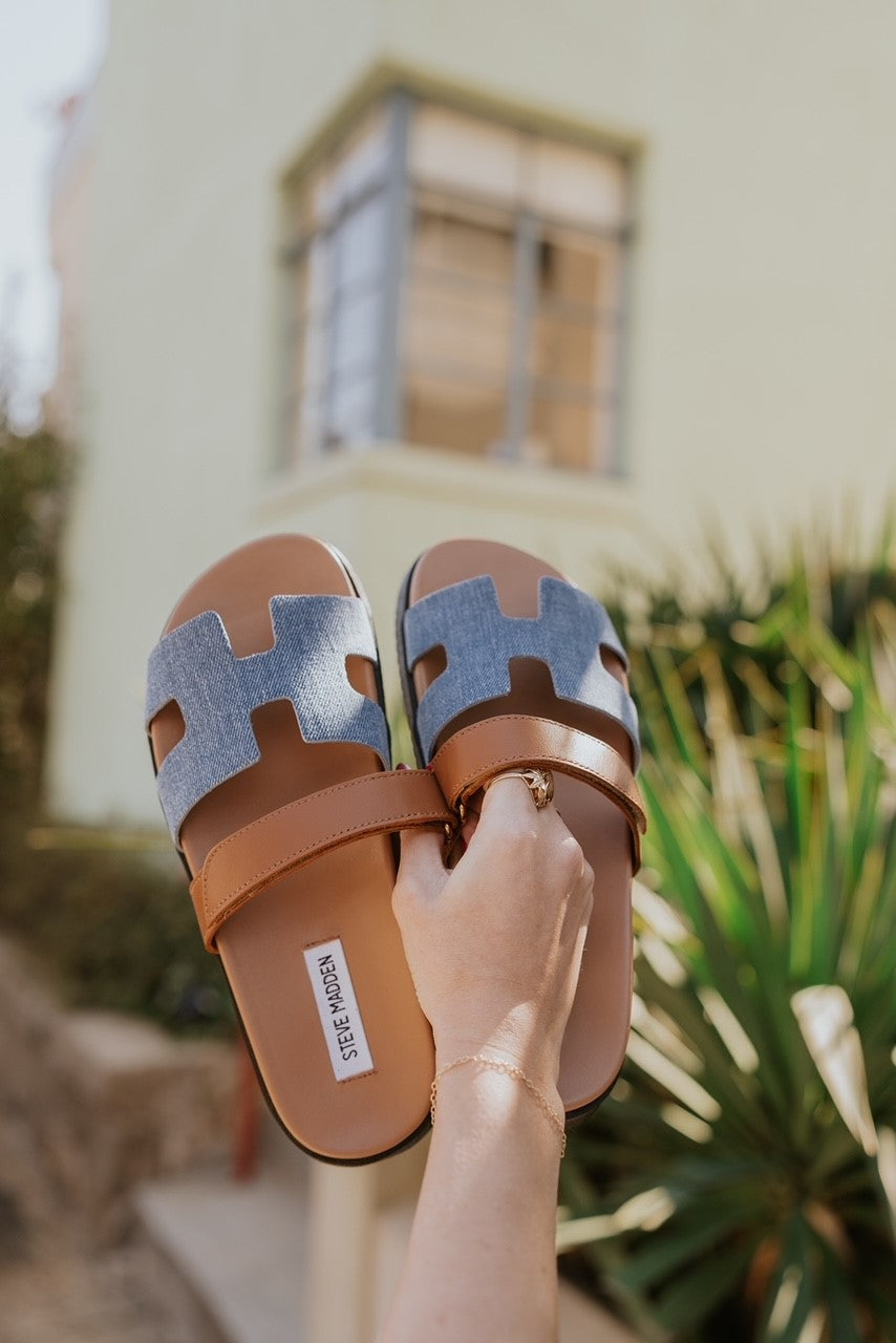 Ariel view of the model holding the Mayven Sandal in Denim Multi which features a brown leather fabric cutout strappy upper with hook and loop closure, denim "h" shaped strap, round toe, black sole and slide on style.
