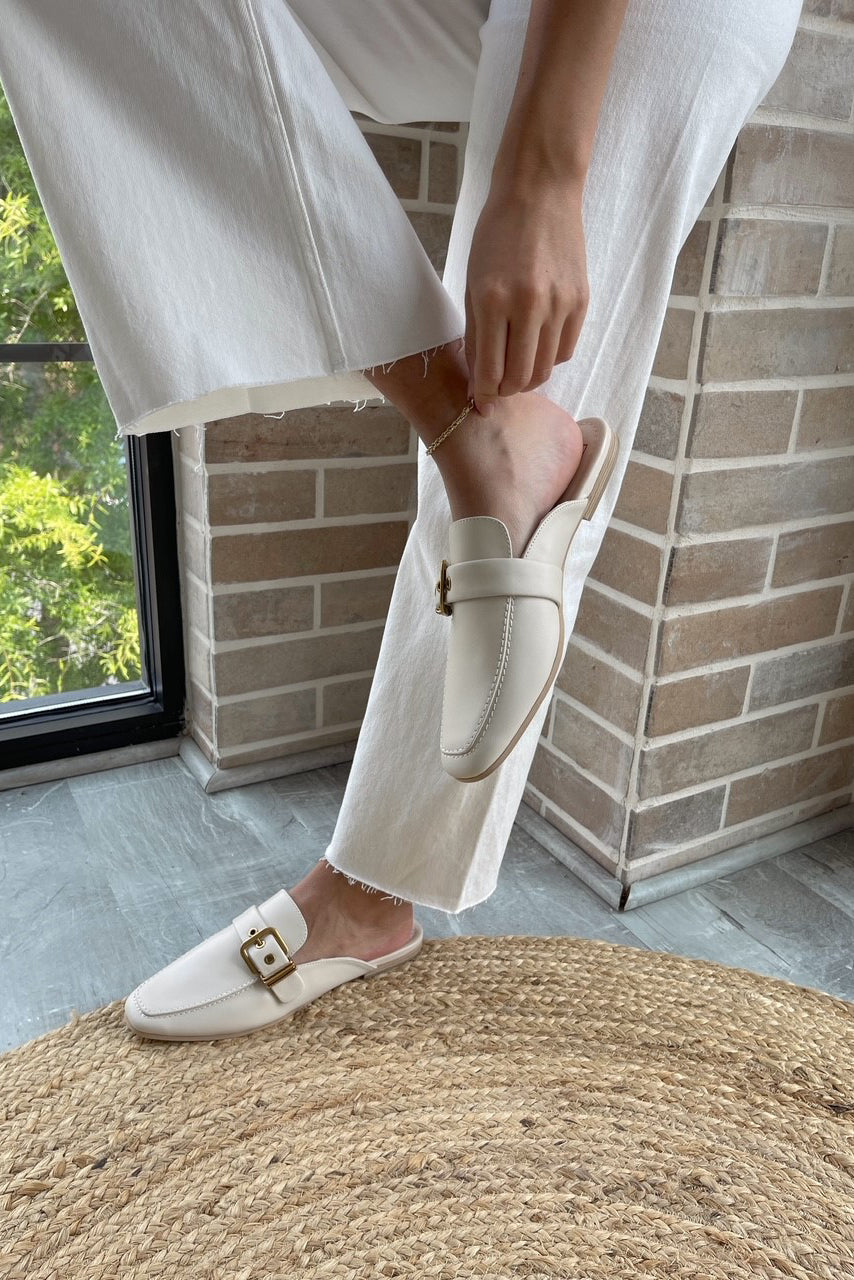 Front angle view of female model wearing the Santel Flat in Ivory Leather which features ivory leather fabric, square round toe, slide-on style, gold adjustable buckle detail and flat loafer.