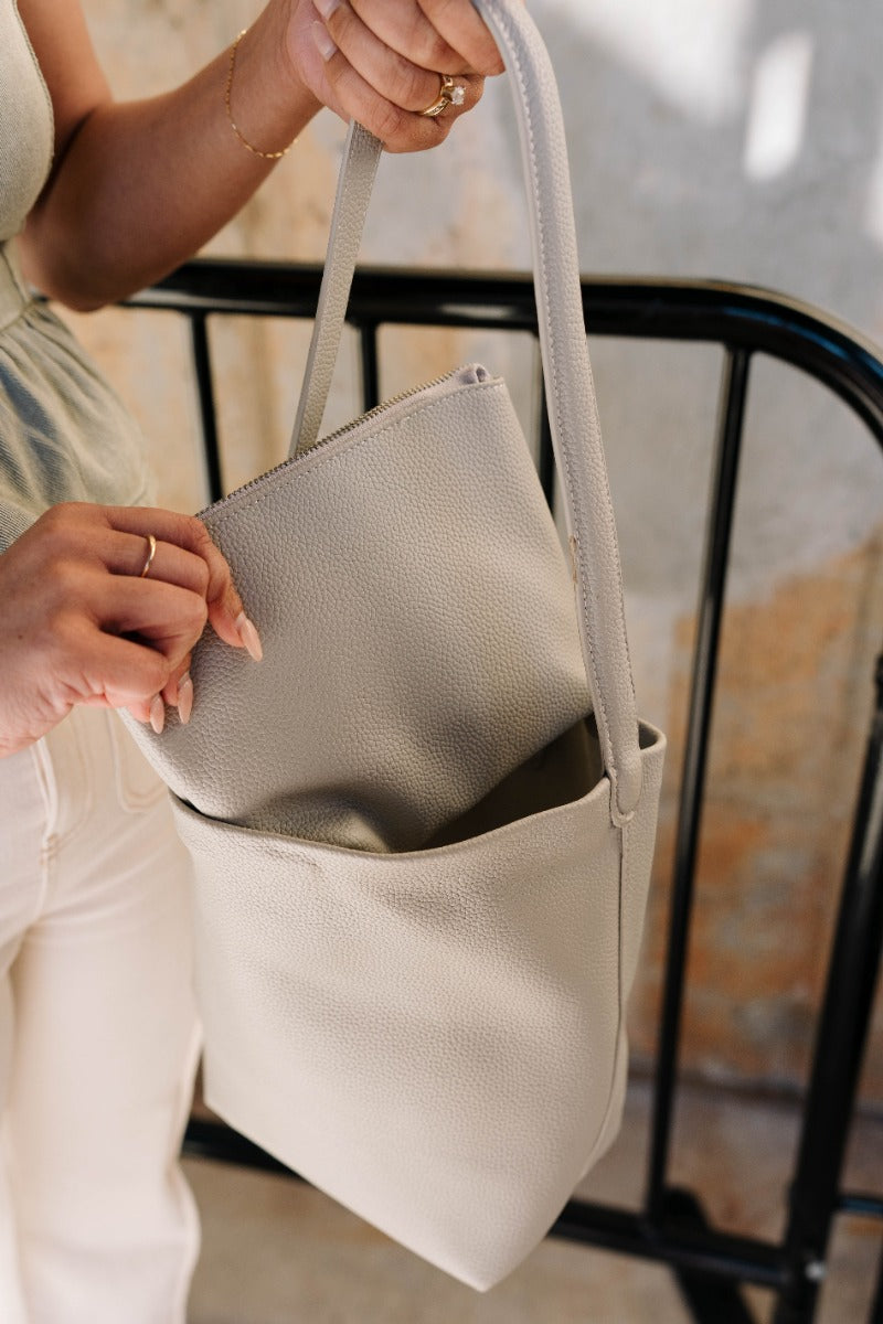 Model is holding the Belinda Grey Faux-Leather Purse that has stone grey faux leather fabric, two shoulder straps, cream lining, a monochrome interior tote and a button snap closure.