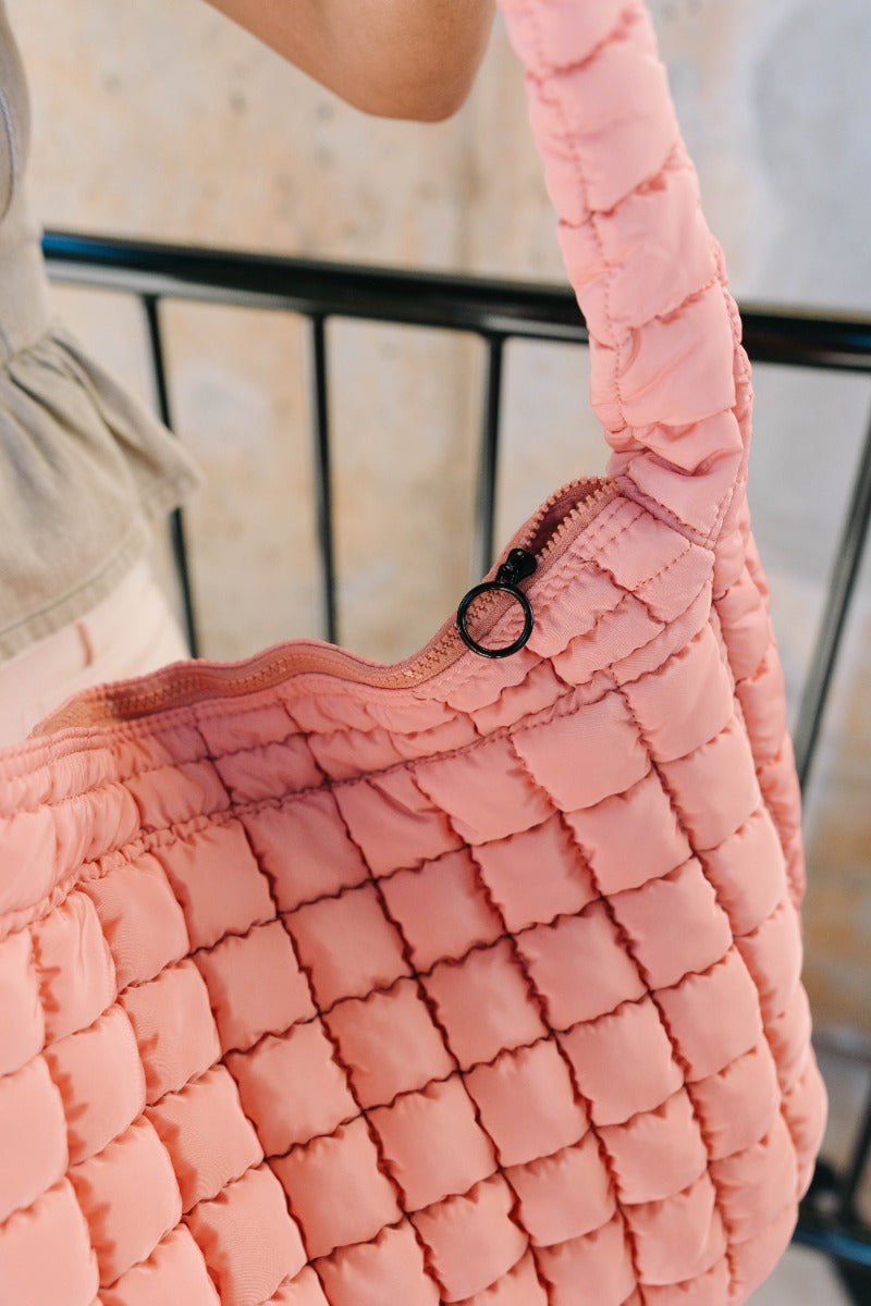 Close up view of model holding the Sedona Blush Puffer Oversized Tote which features blush pink puffer fabric, two front pockets with monochrome zipper closure, one large moncohrome zipper, pockets on the inside and a single strap.