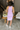 Full body back view of model wearing the Out And About Dress In Lavender which features lavender knit fabric, ruched side details, midi length, lavender lining, a round neckline and a sleeveless design.