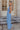 Full body side view of model wearing the Everly Chambray Blue Halter Neckline Jumpsuit which features light blue lightweight fabric, pleated details, two front slit pockets, wide pant legs, light blue shorts lining, a halter neckline with a twist detail,