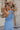 Back view of model wearing the Everly Chambray Blue Halter Neckline Jumpsuit which features light blue lightweight fabric, pleated details, two front slit pockets, wide pant legs, light blue shorts lining, a halter neckline with a twist detail, a sleevele